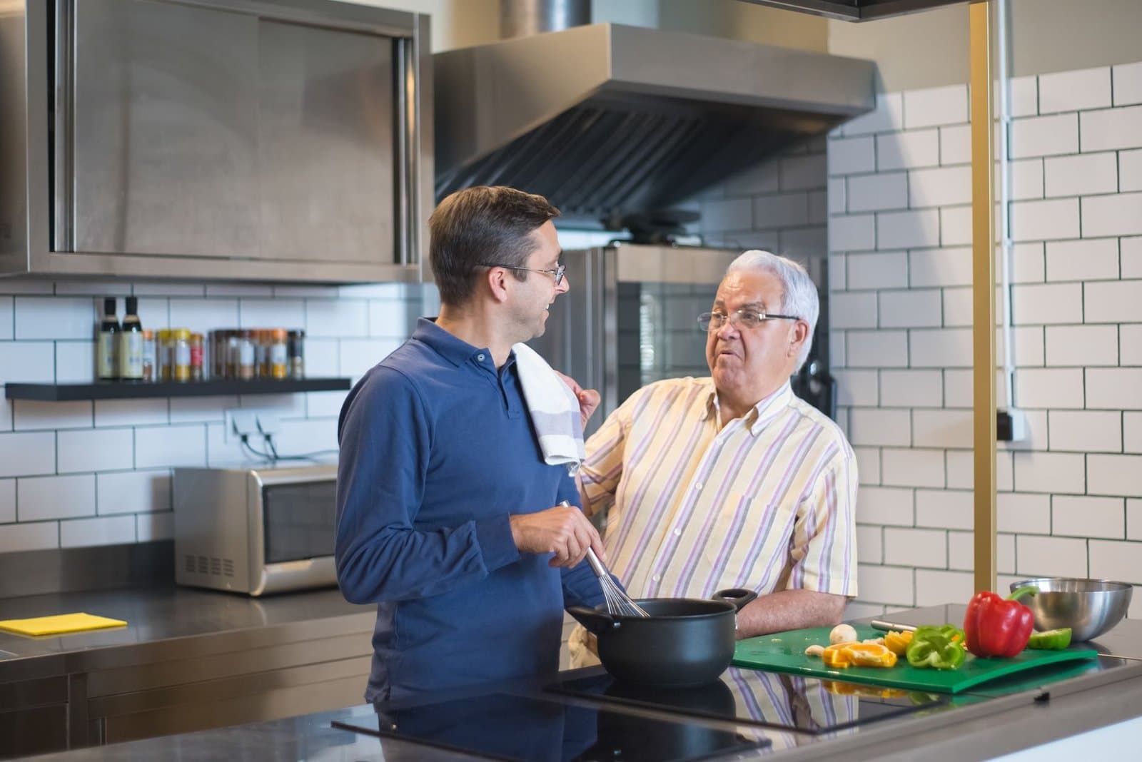 Pai idoso e filho adulto conversando na cozinha enquanto preparam uma refeicao juntos. Foto de Kampus Production via Pexels.